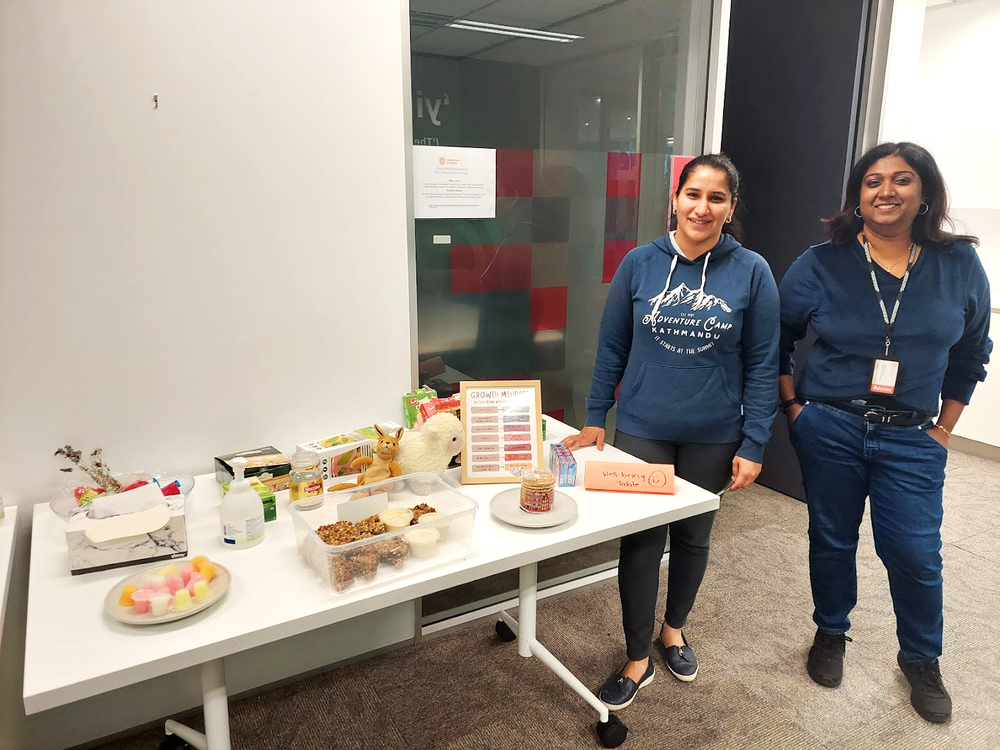 Two students standing next to a table displaying sensory items shared for wellbeing. Items include lavender flowers, soft toys, instant coffee, herbal teas, and biscuits.