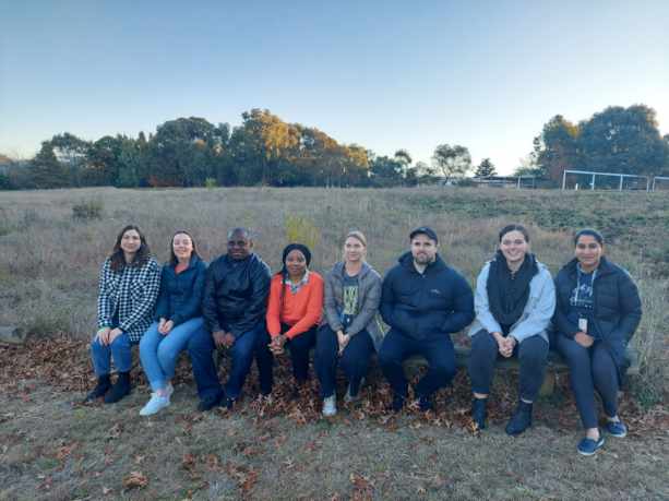 Group work class sitting in a row in front of an open grass field with trees on the horizon behind them. The image illustrates a group bonding during a walk and talk activity.